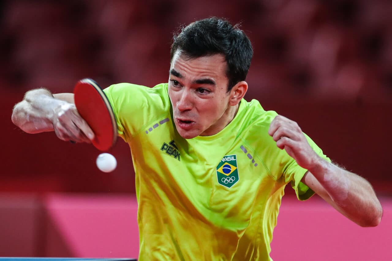 Hugo Calderano é branco, usa uma camiseta amarela e segura uma raquete de tênis de mesa, ele acerta uma bolinha de tênis de mesa branca.