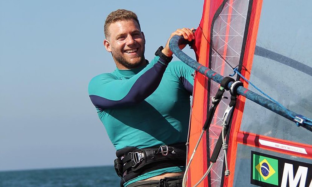 Mateus isaac, atleta da classe iQFoil da vela, sorri enquanto segura sua embarcação na Semana Olímpica Francesa de Hyères