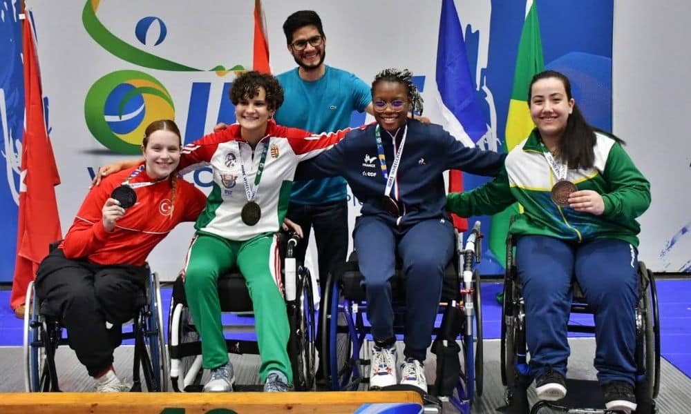 Ana Elisa posa para foto com as medalhistas. As quatro estão em suas cadeiras de rodas. Ao fundo, um homem barbado posa com elas, usando camisa azul