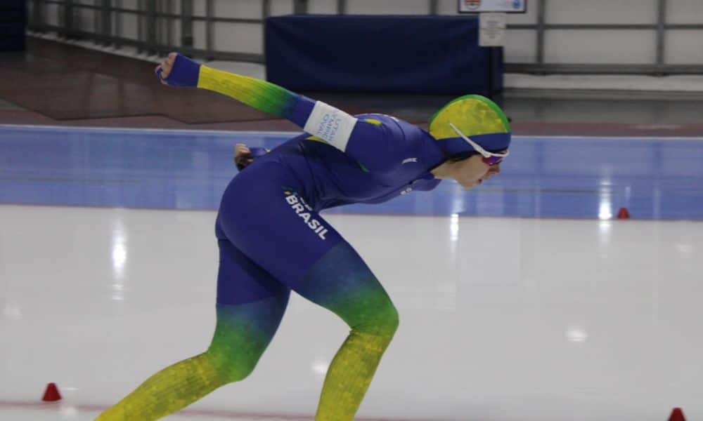 Larissa Paes patina sobre o gelo. Ela veste uma malha azul com detalhes em amarelo e um par de óculos de proteção