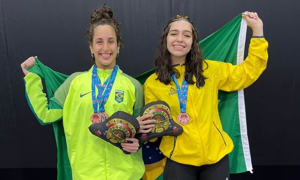 Lourdes e Laila posam para foto. Elas vestem uniforme do Time Brasil e estão com as medalhas no peito. Ambas seguram uma bandeira nas costas