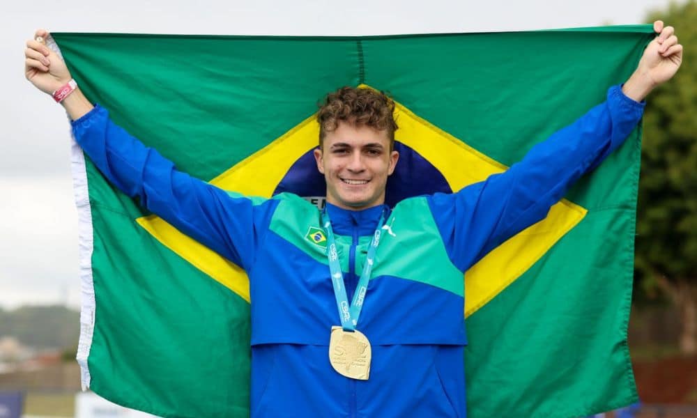 Renan Galina com a medalha de ouro no peito. Ele veste um agasalho azul e verde e tem uma bandeira do Brasil nos braços