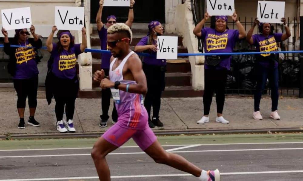 Daniel do Nascimento corre na Maratona de Nova York. Ao fundo, mulheres com cartazes incentivando as pessoas a votar na eleição legislativa dos EUA
