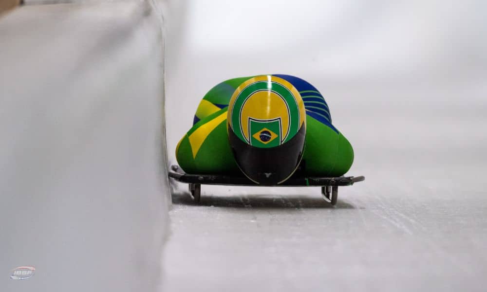 Nicole Silveira deitada no trenó. Ela usa um capacete verde e amarelo com a bandeira do Brasil