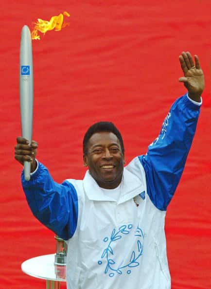 Pelé veste um casaco branco com mangas azuis e segura a tocha de 2004