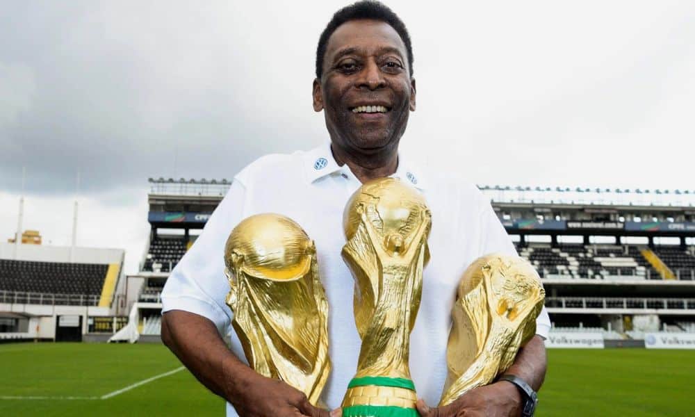Pelé posa para foto na Vila Belmiro. Ele veste uma camisa branca e segura três troféus da Copa do Mundo