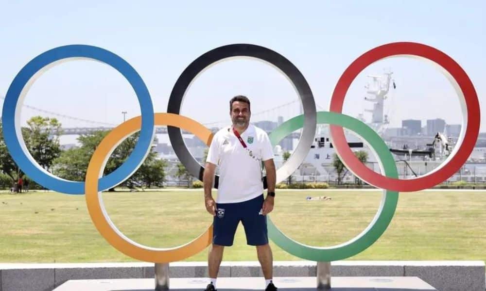 Duda Musa, vestindo bermuda azul e camisa polo branca, posa para foto na frente dos aros olímpicos