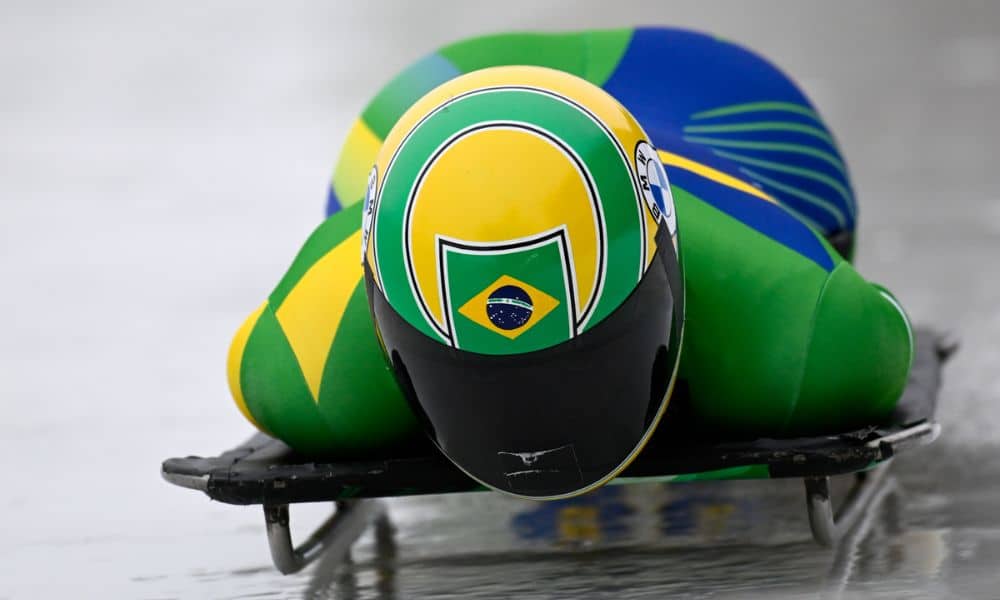 Close de Nicole Silveira no trenó. Ela veste malha verde e capacete amarelo com detalhes em verde, similar ao de Ayrton Senna
