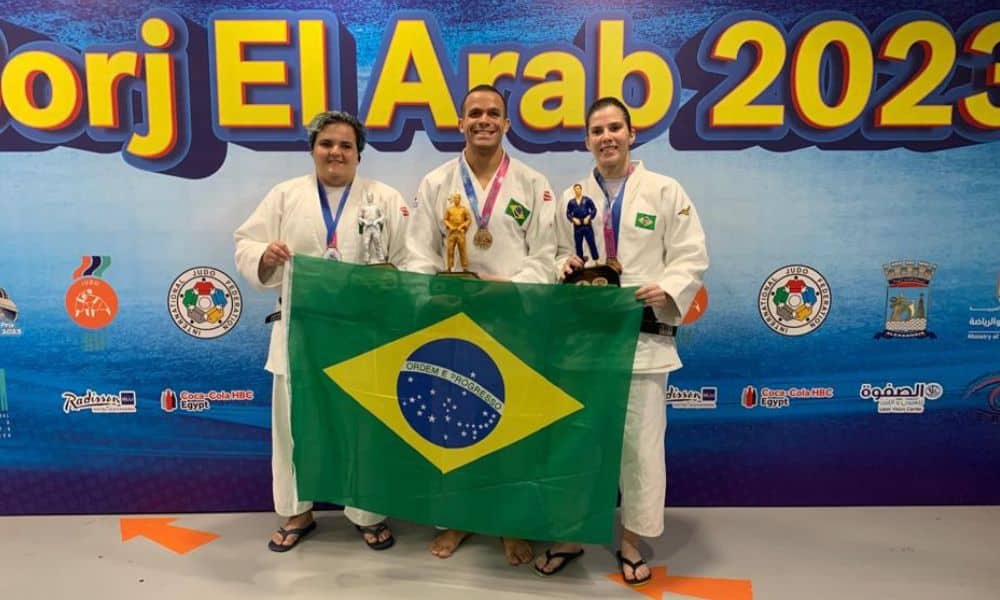 Mas Rebeca, Arthur Silva e Alana posam para foto com suas medalhas no peito. Eles vestem quimono branco e seguram uma bandeira do Brasil mas