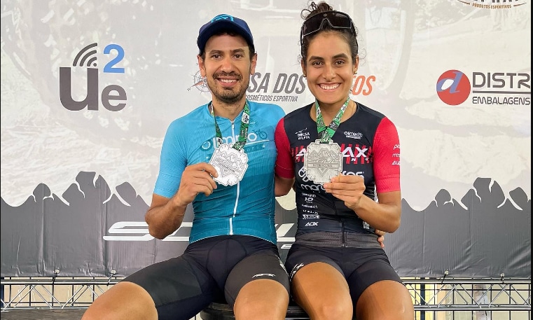 Na imagem, Karen e Rodrigo sentados no lugar mais alto do pódio mostrando as medalhas de vencedores.