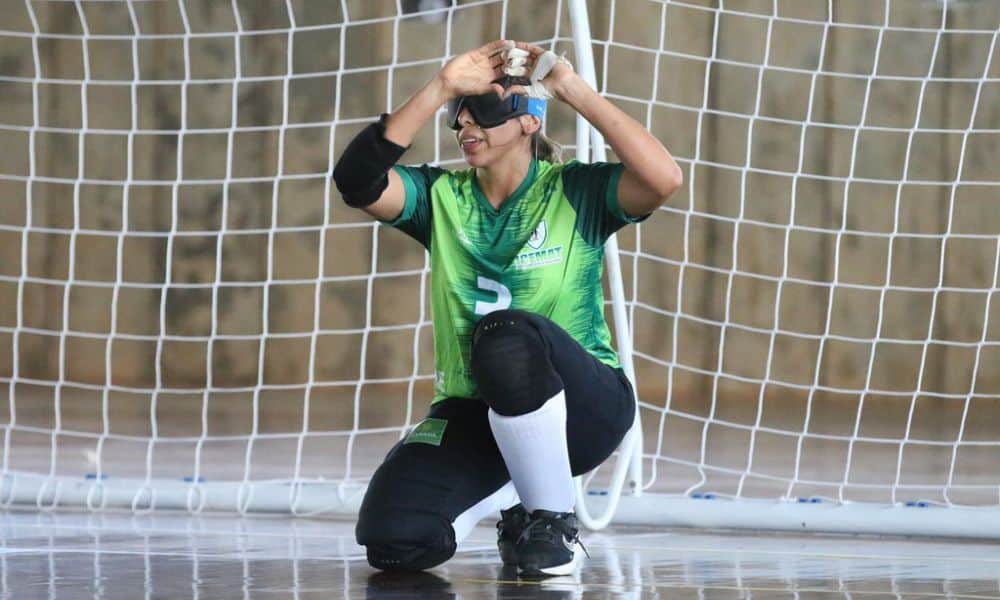 Com o joelho direito apoiado na quadra, Carol Duarte sorri e une as duas mãos à frente do rosto para formar um coração, na comemoração de seu gol na semifinal do Regional Centro-Norte de goalball (Foto: Renan Cacioli/ CBDV)