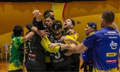 Jogadores e comissão técnica do Athlon se abraçam, emocionados, em comemoração à vitória no Brasileiro de goalball. Eles estão na quadra e há um fundo amarelo (Foto: Taba Benedicto/CBDV)
