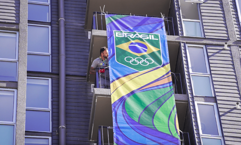 Trabalhador está na sacada de um prédio na Vila Pan-Americana ajustando uma plotagem com a marca do Time Brasil