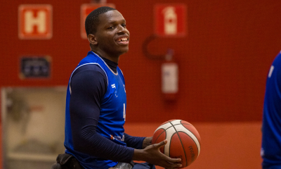 Jogador da seleção brasileira de basquete em cadeira de rodas está de camiseta azul, com a bola na mão, e sorri. Ao fundo, há uma parede vermelha. Ele estará nos Jogos Parapan-Americanos de Santiago-2023.