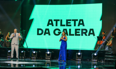 Jornalistas Everaldo Marques (terno cinza) e Karine Alves (vestido azul) seguram o microfone e aparecem no centro da imagem, em cima do palco apresentando o Prêmio Paralímpicos 2022. Ao fundo, há uma tela verde com os dizeres "Atleta da Galera"