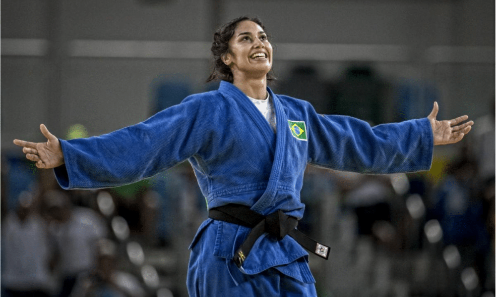 Mariana Silva usa quimono azul enquanto sorri de braços abertos após luta de judô