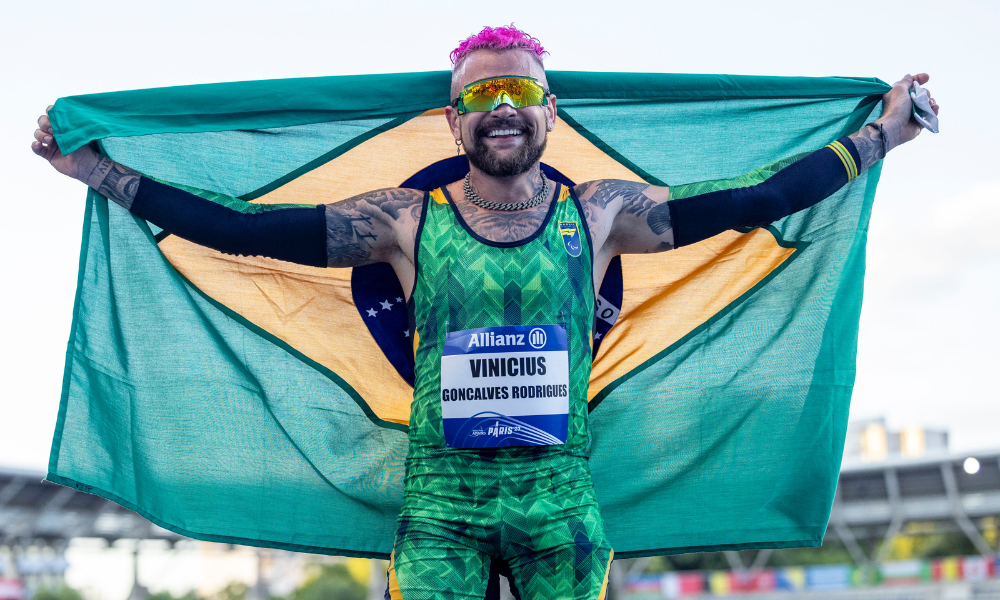 Vinicius Rodrigues com a bandeira do Brasil enquanto sorri para foto; ele está no BBB 24 e teve o pagamento do Bolsa Atleta suspenso