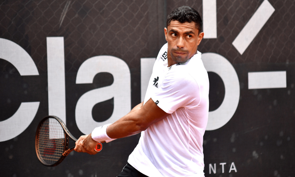 Thiago Monteiro está com a raquete na mão em movimento de ataque em frente ao backdrop do Rio Open; ele enfrentará Carlos Alcaraz na primeira rodada