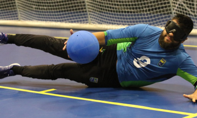 André Dantas está na ala esquerda e cai para o lado na defesa da bola, que atinge a altura de suas pernas, durante treino. Ele veste calça preta e camisa de manga longa azul com detalhes em verde. Foto: Renan Cacioli/ CBDV.