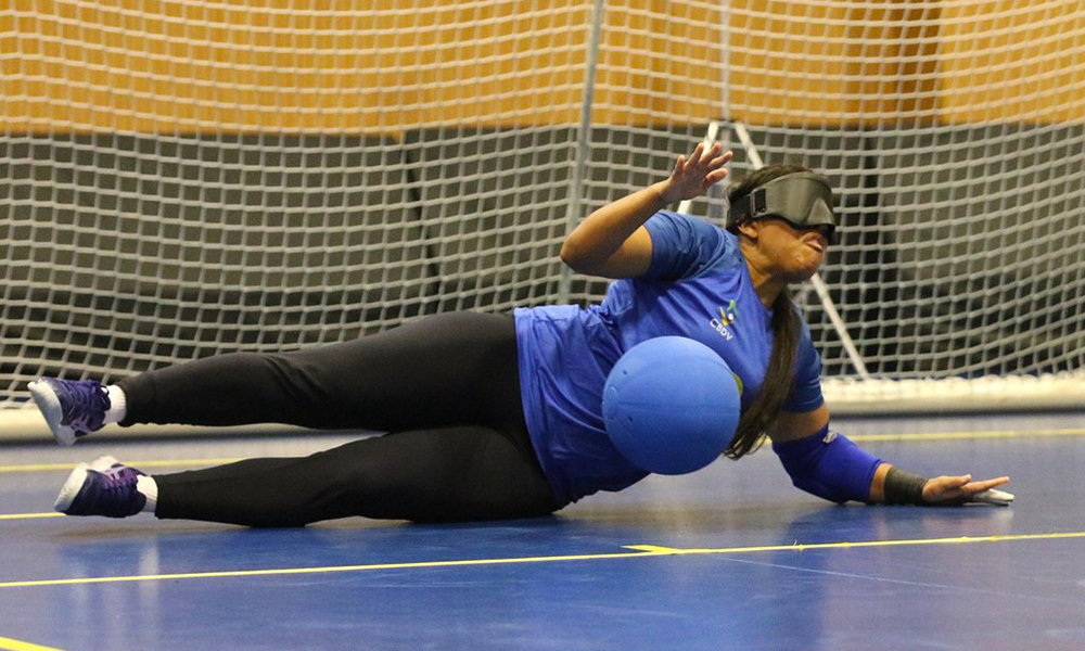 Na imagem, Jéssica Vitorino, da Seleção Brasileira, tem as pernas bem esticadas e apoia o peso do corpo no chão da quadra com sua mão esquerda enquanto executa o movimento de defesa da bola, que se aproxima do seu tronco.
