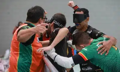 Na imagem, Grupo da AGAFUC comemora na banda lateral. Dois atletas, Gabriel e Nonato, usam máscaras pretas para os olhos e protetores bucais. Os jogadores se abraçam e parecem muito felizes. De camiseta laranja e verde, o técnico Rafael Astrada estende a mão, enquanto o goleiro Luan, de uniforme verde, está curvado no abraço coletivo. O fundo é um ginásio esportivo coberto com uma parede de tijolos cinza.