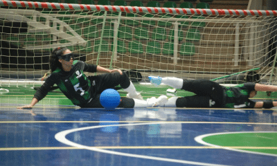 Quadra esportiva com duas jogadoras do ICEMAT deitadas no chão, próximas à linha do gol. Ambas usam vendas nos olhos e uniforme preto com listras verdes. Uma delas, com o número 5, está sentada com uma das mãos apoiada no chão e a outra perna dobrada, enquanto a bola azul está encostada em sua perna. A outra jogadora está totalmente deitada, com os braços esticados para frente, como se estivesse tentando defender a bola. Ao fundo, há uma trave de gol e arquibancadas vazias com cadeiras verdes