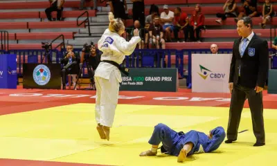 Rosi Andrade, de quimono branco, está saltando com ambos os pés para fora do chão, aparentemente comemorando. No chão, uma oponente de quimono azul está deitada de costas