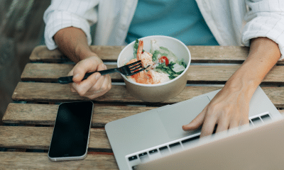 Pessoa trabalhando com notebook, celular e comida ao lado, representando refeição feita no automático em meio ao acúmulo de tarefas.