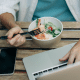Pessoa trabalhando com notebook, celular e comida ao lado, representando refeição feita no automático em meio ao acúmulo de tarefas.