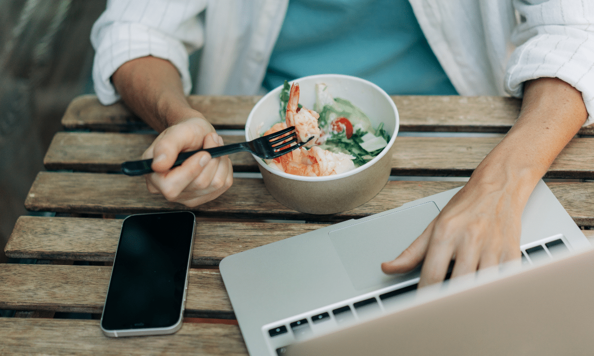 Pessoa trabalhando com notebook, celular e comida ao lado, representando refeição feita no automático em meio ao acúmulo de tarefas.