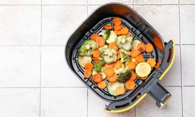 Foto horizontal de uma air fryer aberta ou cesta sobre uma bancada com legumes coloridos já preparados ou prontos para assar, como abobrinha, cenoura, brócolis, couve-flor e batata-doce