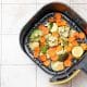 Foto horizontal de uma air fryer aberta ou cesta sobre uma bancada com legumes coloridos já preparados ou prontos para assar, como abobrinha, cenoura, brócolis, couve-flor e batata-doce