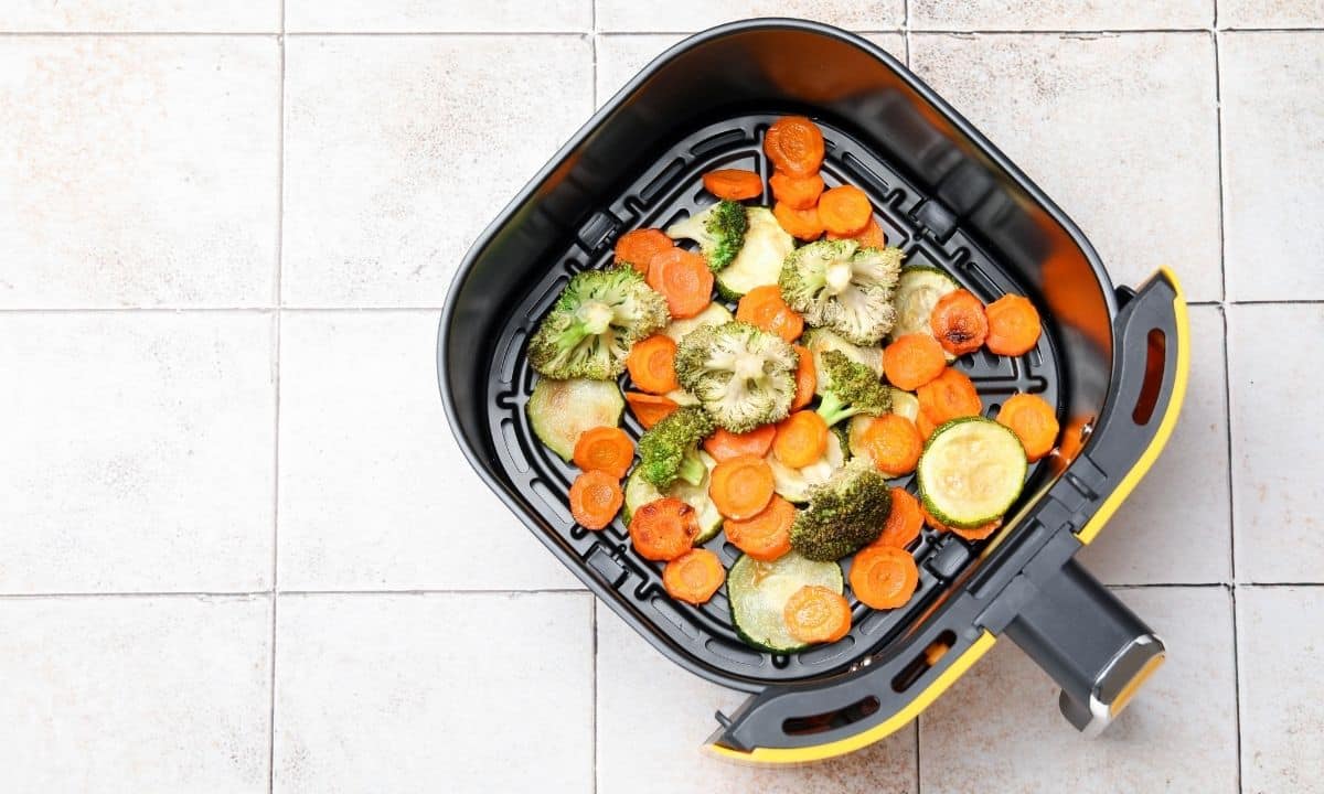 Foto horizontal de uma air fryer aberta ou cesta sobre uma bancada com legumes coloridos já preparados ou prontos para assar, como abobrinha, cenoura, brócolis, couve-flor e batata-doce