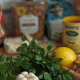 Foto horizontal de uma bancada de cozinha com alimentos embalados ao fundo e, em primeiro plano, ervas frescas, limão, alho, cebola e especiarias.