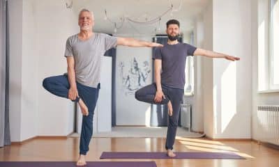 Foto horizontal de uma pessoa em pé, próxima a uma parede ou cadeira, fazendo exercício de equilíbrio em um pé só