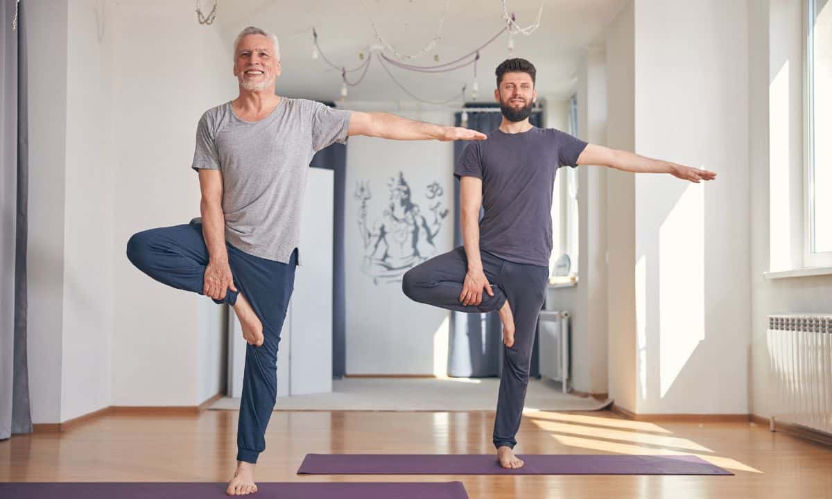 Foto horizontal de uma pessoa em pé, próxima a uma parede ou cadeira, fazendo exercício de equilíbrio em um pé só