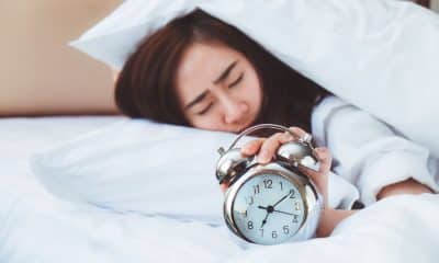 pessoa acordando com dificuldade em uma manhã de segunda-feira, com despertador ao lado da cama e luz entrando pela janela.