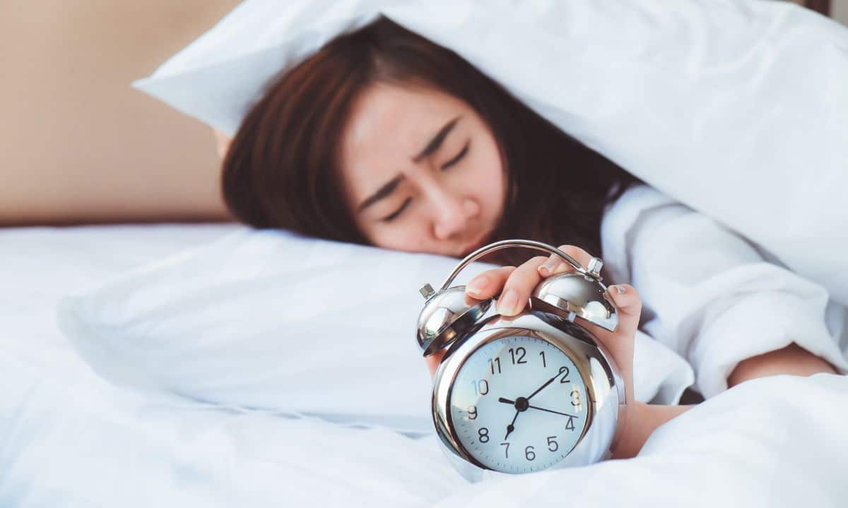 pessoa acordando com dificuldade em uma manhã de segunda-feira, com despertador ao lado da cama e luz entrando pela janela.