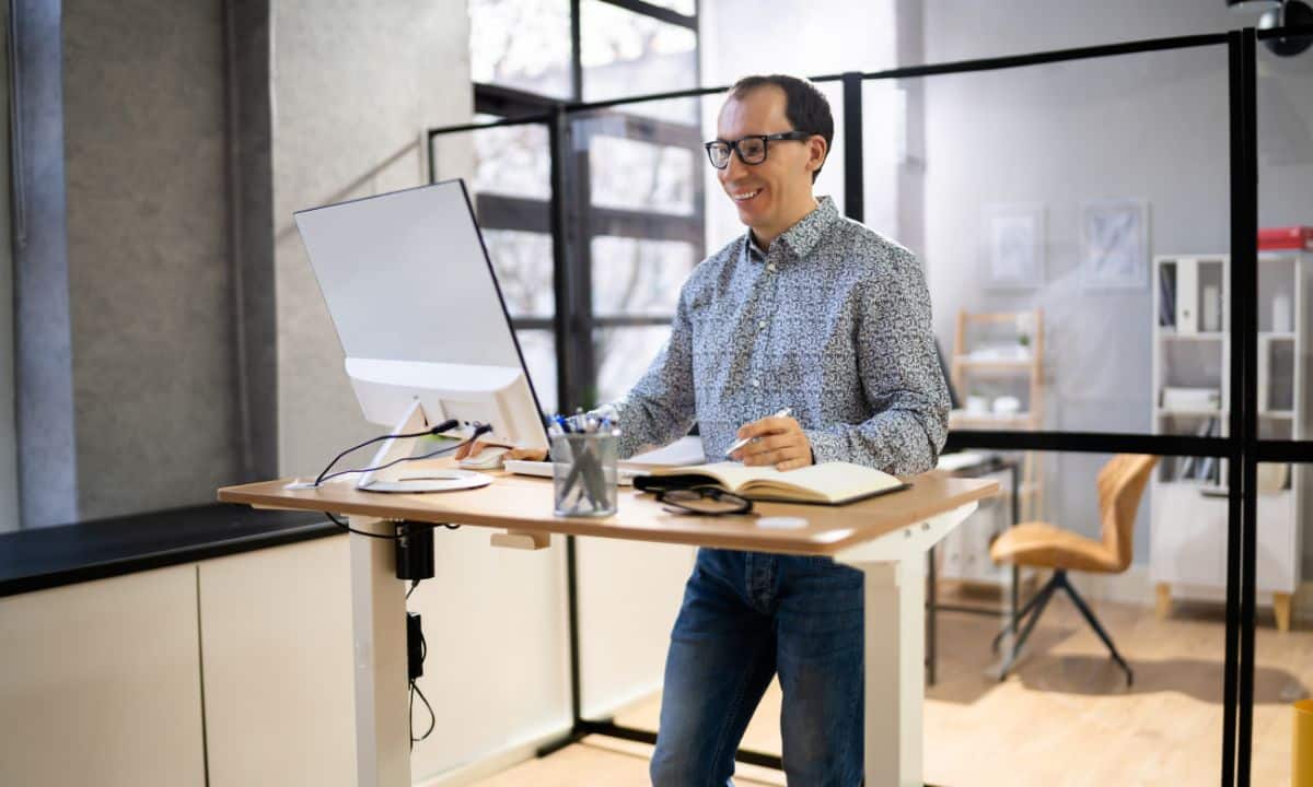 pessoa em home office ou escritório participando de reunião no notebook, em pé ao lado da mesa ou fazendo ajuste discreto de postura.