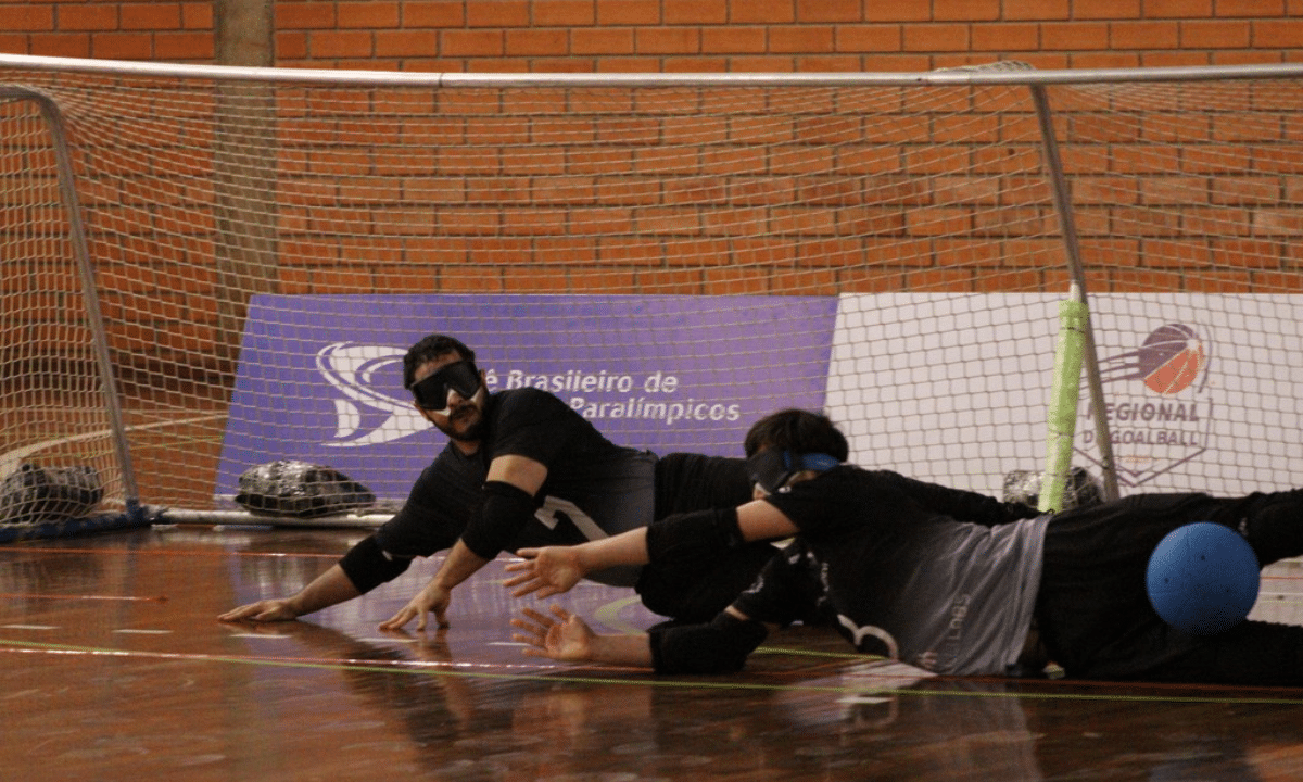 Dois atletas de goalball, usando vendas nos olhos e uniforme preto e cinza, estão deitados em frente ao gol durante uma jogada defensiva. Ao fundo, aparecem as redes da baliza e banners da competição.