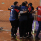 Grupo de jogadores de goalball do IRM comemorando abraçados em frente ao gol dentro de um ginásio. Todos vestem uniforme azul com detalhes em vermelho e preto; alguns jogadores usam vendas esportivas nos olhos. No centro do grupo, os atletas estão bem próximos, com braços pelos ombros uns dos outros, transmitindo sensação de celebração ou união após uma jogada. À direita, um integrante da comissão técnica, com colete vermelho escrito “COMISSÃO TÉCNICA” e “ACESA” nas costas, toca o ombro de um dos jogadores, participando do momento. O gol de goalball, com rede branca, ocupa quase toda a largura da imagem, e bolas de goalball prateadas com faixas pretas estão encostadas na base da trave. Crédito: Alejandro Aisi/CBDV