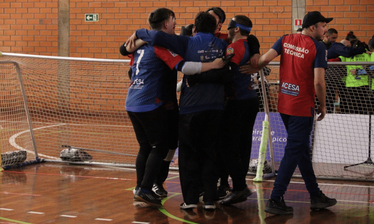Grupo de jogadores de goalball do IRM comemorando abraçados em frente ao gol dentro de um ginásio. Todos vestem uniforme azul com detalhes em vermelho e preto; alguns jogadores usam vendas esportivas nos olhos. No centro do grupo, os atletas estão bem próximos, com braços pelos ombros uns dos outros, transmitindo sensação de celebração ou união após uma jogada. À direita, um integrante da comissão técnica, com colete vermelho escrito “COMISSÃO TÉCNICA” e “ACESA” nas costas, toca o ombro de um dos jogadores, participando do momento. O gol de goalball, com rede branca, ocupa quase toda a largura da imagem, e bolas de goalball prateadas com faixas pretas estão encostadas na base da trave. Crédito: Alejandro Aisi/CBDV