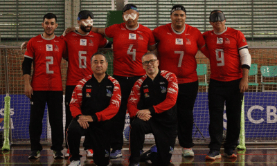 Grupo da IRM posando em uma quadra esportiva de piso brilhante, em frente a uma rede de goalball. Ao fundo há cinco jogadores em pé, todos com camisa vermelha e calça preta; na frente há dois homens ajoelhados, usando agasalhos esportivos pretos com mangas vermelhas. Os cinco atletas de trás vestem camisas vermelhas com detalhes brancos nas mangas e golas, número grande prateado no centro e vários logotipos pequenos na parte superior do peito. Da esquerda para a direita, os números nas camisas são: 2, 6, 4, 7 e 9. Três dos jogadores usam vendas ou óculos escuros de proteção próprios de goalball; o jogador com número 6 tem ataduras brancas sobre os olhos sob o óculos. Crédito: Alejandro Aisi/CBDV