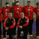 Grupo da IRM posando em uma quadra esportiva de piso brilhante, em frente a uma rede de goalball. Ao fundo há cinco jogadores em pé, todos com camisa vermelha e calça preta; na frente há dois homens ajoelhados, usando agasalhos esportivos pretos com mangas vermelhas. Os cinco atletas de trás vestem camisas vermelhas com detalhes brancos nas mangas e golas, número grande prateado no centro e vários logotipos pequenos na parte superior do peito. Da esquerda para a direita, os números nas camisas são: 2, 6, 4, 7 e 9. Três dos jogadores usam vendas ou óculos escuros de proteção próprios de goalball; o jogador com número 6 tem ataduras brancas sobre os olhos sob o óculos. Crédito: Alejandro Aisi/CBDV
