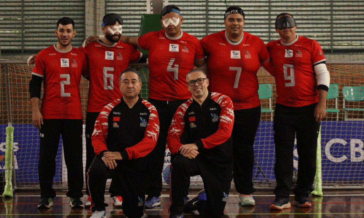 Grupo da IRM posando em uma quadra esportiva de piso brilhante, em frente a uma rede de goalball. Ao fundo há cinco jogadores em pé, todos com camisa vermelha e calça preta; na frente há dois homens ajoelhados, usando agasalhos esportivos pretos com mangas vermelhas. Os cinco atletas de trás vestem camisas vermelhas com detalhes brancos nas mangas e golas, número grande prateado no centro e vários logotipos pequenos na parte superior do peito. Da esquerda para a direita, os números nas camisas são: 2, 6, 4, 7 e 9. Três dos jogadores usam vendas ou óculos escuros de proteção próprios de goalball; o jogador com número 6 tem ataduras brancas sobre os olhos sob o óculos. Crédito: Alejandro Aisi/CBDV