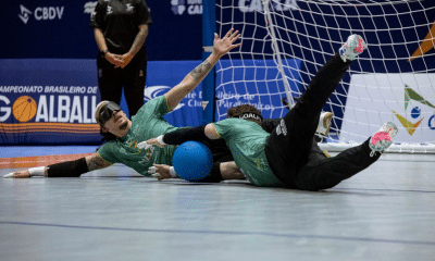 Dois atletas do ATHLON estão deitados no chão da quadra, próximos à trave, durante uma jogada defensiva. Ambos usam uniformes verdes, calças pretas e vendas nos olhos. Um deles está estendido de lado, com um braço levantado, enquanto o outro está com as pernas erguidas, tentando defender a bola azul, que está entre os dois. Crédito: Taba Benedicto/CBDV