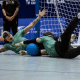 Dois atletas do ATHLON estão deitados no chão da quadra, próximos à trave, durante uma jogada defensiva. Ambos usam uniformes verdes, calças pretas e vendas nos olhos. Um deles está estendido de lado, com um braço levantado, enquanto o outro está com as pernas erguidas, tentando defender a bola azul, que está entre os dois. Crédito: Taba Benedicto/CBDV
