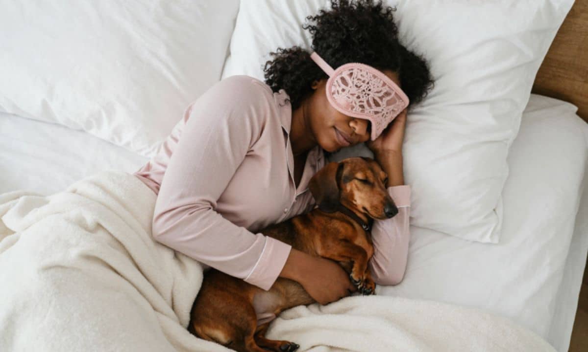 Pessoa descansa na cama enquanto o cachorro dorme ao seu lado.