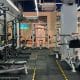 Foto horizontal mostrando, no mesmo enquadramento, uma máquina de musculação e um suporte com halteres