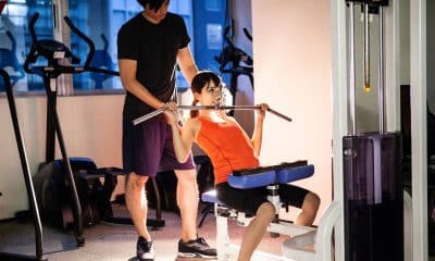 foto horizontal de uma pessoa adulta iniciante recebendo orientação de um professor em uma máquina de musculação. O ambiente pode ter outros frequentadores ao fundo